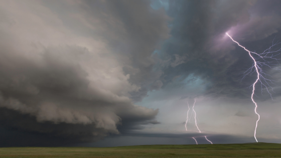 A green field with dark grey storm clouds overhead and three cloud-to-ground lightning strikes