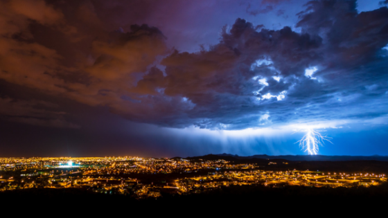 A lit up city view taken from a high elevation with a thunderstorm overhead, complete with bright lightning strikes